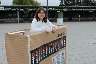 Eine Aktivistin in einem weißen Maleranzug steht in dem überdimensionierten Pakte, im Hintergrund ist eine Straßenbahn zu sehen.