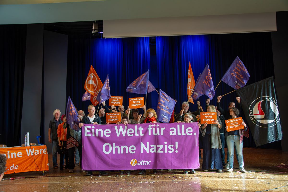 Bild auf dem Attac-Herbstratschlag 2024 in Frankfurt auf dem junges Attac dabei war. Auf dem Bild ist eine Gruppe Menschen zu sehen, die ein Banner mit der Aufschrift "Eine Welt für alle. Ohne Nazis!" halten. Im Hintergrund sind Attac-Flaggen zu sehen.