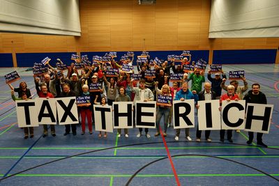 Totalaufnahme der Menschengruppe in der Turnhalle. Die erste Reihe hält A1 große Buchstaben, die "Tax the Rich" bilden, die hinteren kleine Poster auf denen auch "Tax the Rich" steht