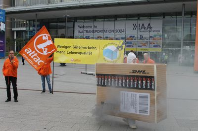 Vor der Messe Hannover stehen Aktivist*innen, die ein Banner halten mit dem Text "Weniger Güterverkehr – mehr Lebensqualität". Davon sieht man ein überdimensioniertes Paket mit einem Auspuff aus dem dunkler Rauch kommt.