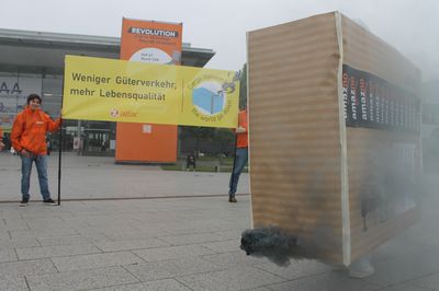 Vor der Messe Hannover stehen Aktivist*innen, die ein Banner halten mit dem Text "Weniger Güterverkehr – mehr Lebensqualität". Davon sieht man ein überdimensioniertes Paket mit einem Auspuff aus dem dunkler Rauch kommt.