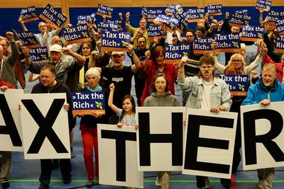 Bild von einer Gruppe von Aktivist*innen die Poster mit dem Slogan "Tax the Rich" hoch halten. Die erste Reihe hält große A1-Buchstaben