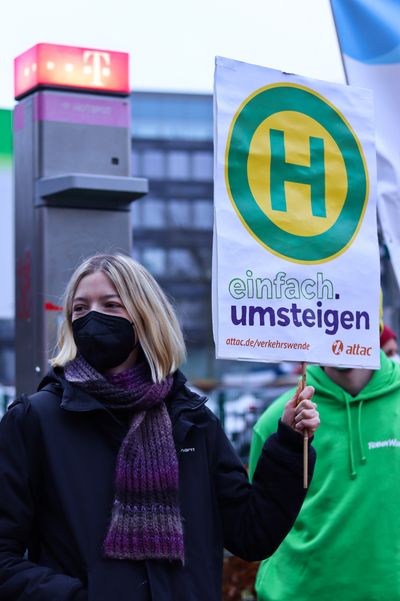 Eine Aktivistin hält ein Schild auf dem ein Haltestellenschilt und der Slogan der Attac-Kampagne "einfach.umsteigen" stehen.