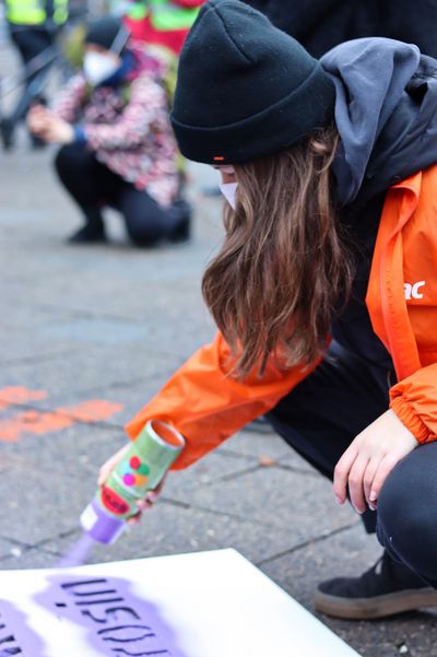 Eine Attac-Aktivistin sprüht Sprühkreide auf eine Schablone die auf dem Gehweg liegt.