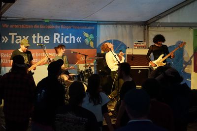 Eine Rockband spielt auf der Bühne, vor ihnen sind die Silhouetten tanzender Menschen zu sehen, hinter ihnen ein Banner mit dem Slogan "Tax the rich"
