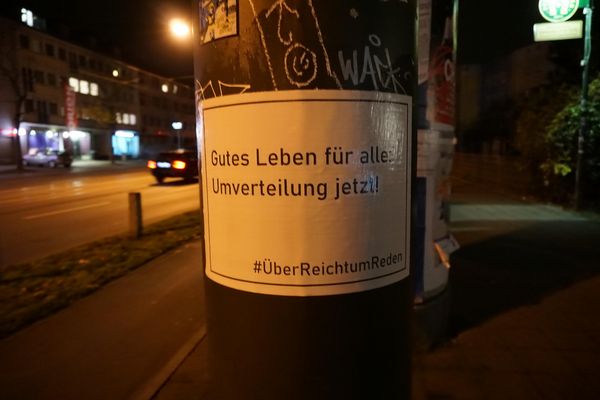 An einem Mast ist ein Plakat mit dem Text "Gutes Leben für alle. Umverteilung jetzt!" angeklebt.