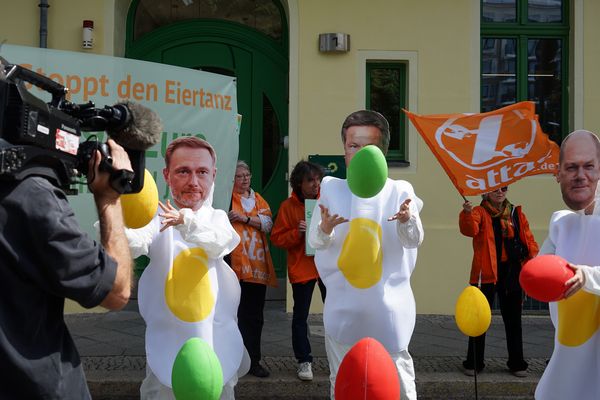 Eine Fernsehkamera nimmt auf, wie drei Aktivist*innen in SPiegeleikostümen mit Masken von Scholz, Habeck und Lindner mit bunten Styroporeiern jonglieren, im Hintergrund sieht man Attacies mit Fahnen und den Eingang der Grünen Parteizentrale