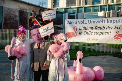 drei Attacies mit Plüschflamingos vor Transparent "Akku voll, Umwelt kaputt - Lithium-Raub der EU in Chile stoppen!" Ein Aktivist trägt eine Habeck-Maske