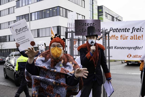 Ein Mensch im Viruskostüm hüpft in erschreckender Mine nach vorne. Im Hintergrund sieht man einen karnevalesk verkleideten Menschen der singt.