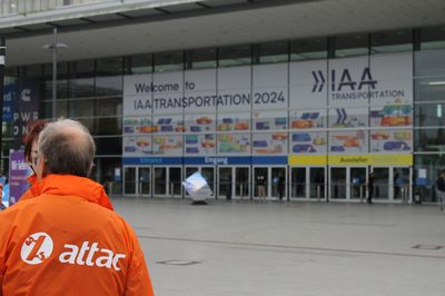 Auf dem Eingang der Messe Hannover ist ein riesiges Plakat mit der Aufschrift "Welcome to IAA-Transportation 2024", davor sieht man von hinten einen Menschen mit einer orangen Jacke auf der groß das Logo von Attac zu sehen ist.